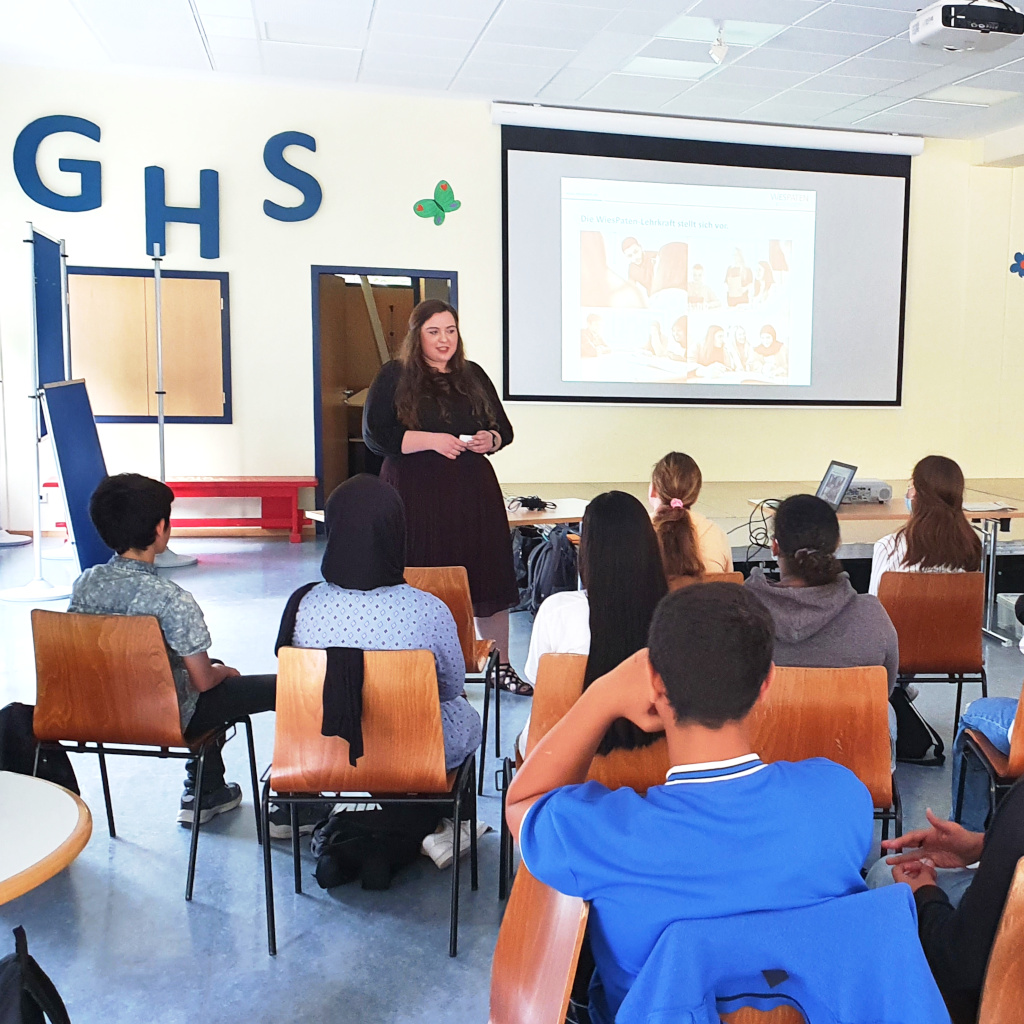 WiesPaten 2022 | Bild von der Infoveranstaltung an der Gerhart-Hauptmann-Schule mit Julius Berger international. Das Bild zeigt Jugendliche in einem Klassenraum. Eine Person hält vorne im Raum an einer Leinwand eine Präsentation.