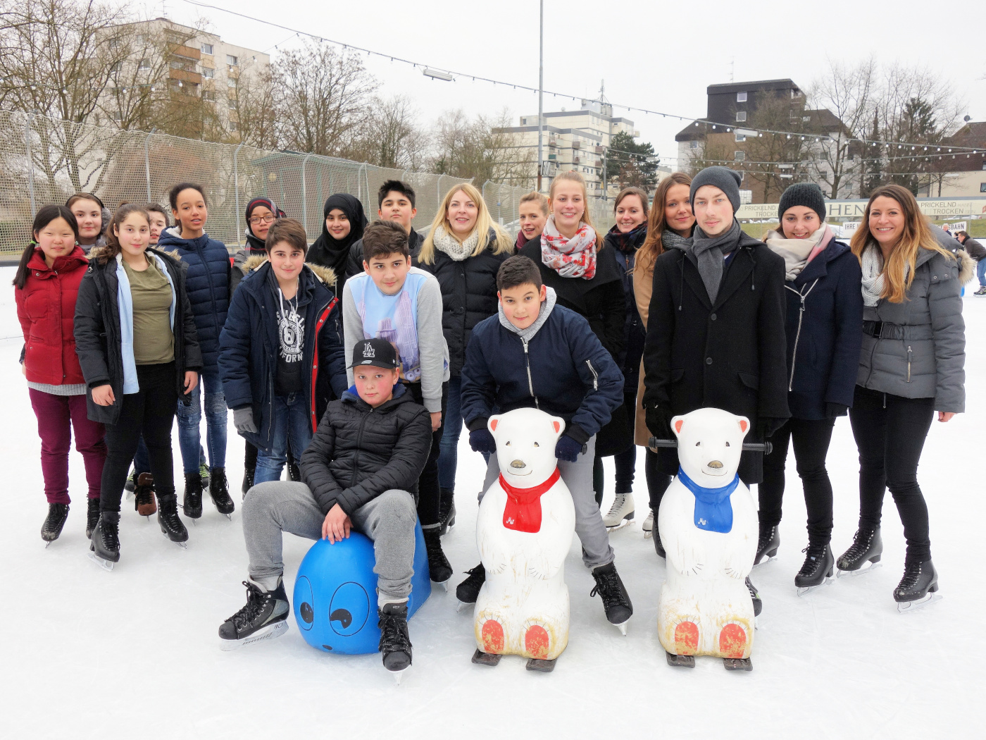wiespaten_event-katalog_henkell-kunsteisbahn Gruppe Jugendlicher stehen mit Patinnen und Paten auf der Eisbahn - im Vordergrund sind "Schiebe-Eisbären" zu sehen, die beim Eislaufen-Lernen unterstützen.